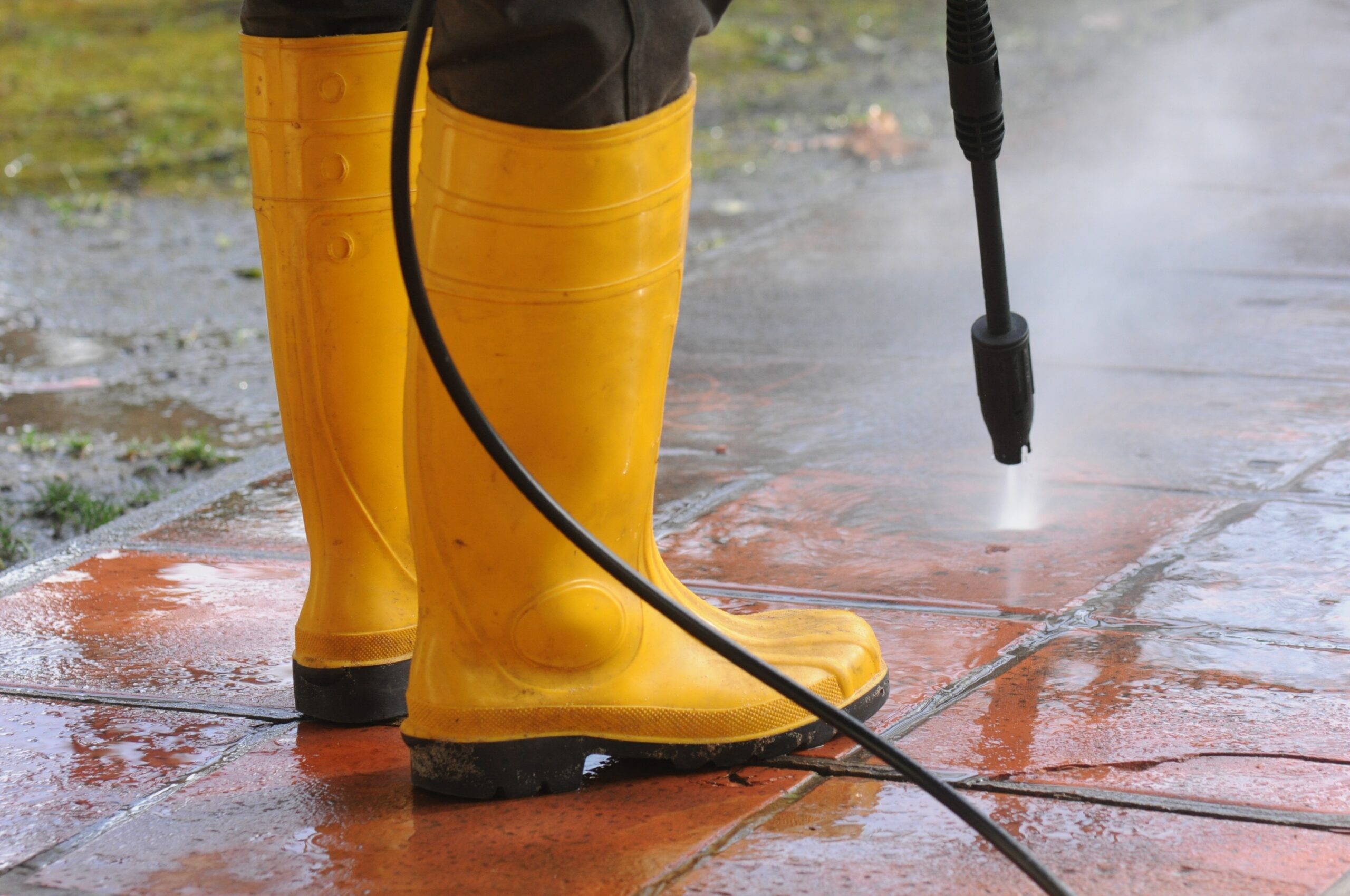 komplekspro.com person wearing yellow rubber boots with high pressure water nozzle cleaning the dirt in the tiles